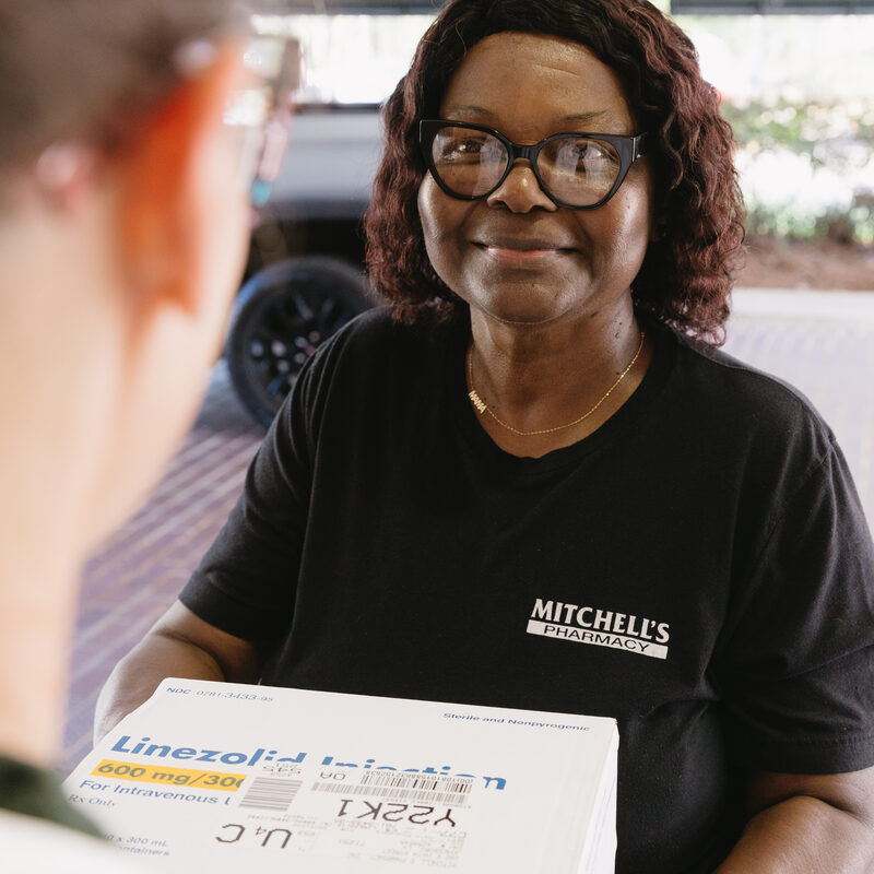 A Mitchell's Pharmacy team member in a Mitchell's Pharmacy shirt handing a medication box to a patient